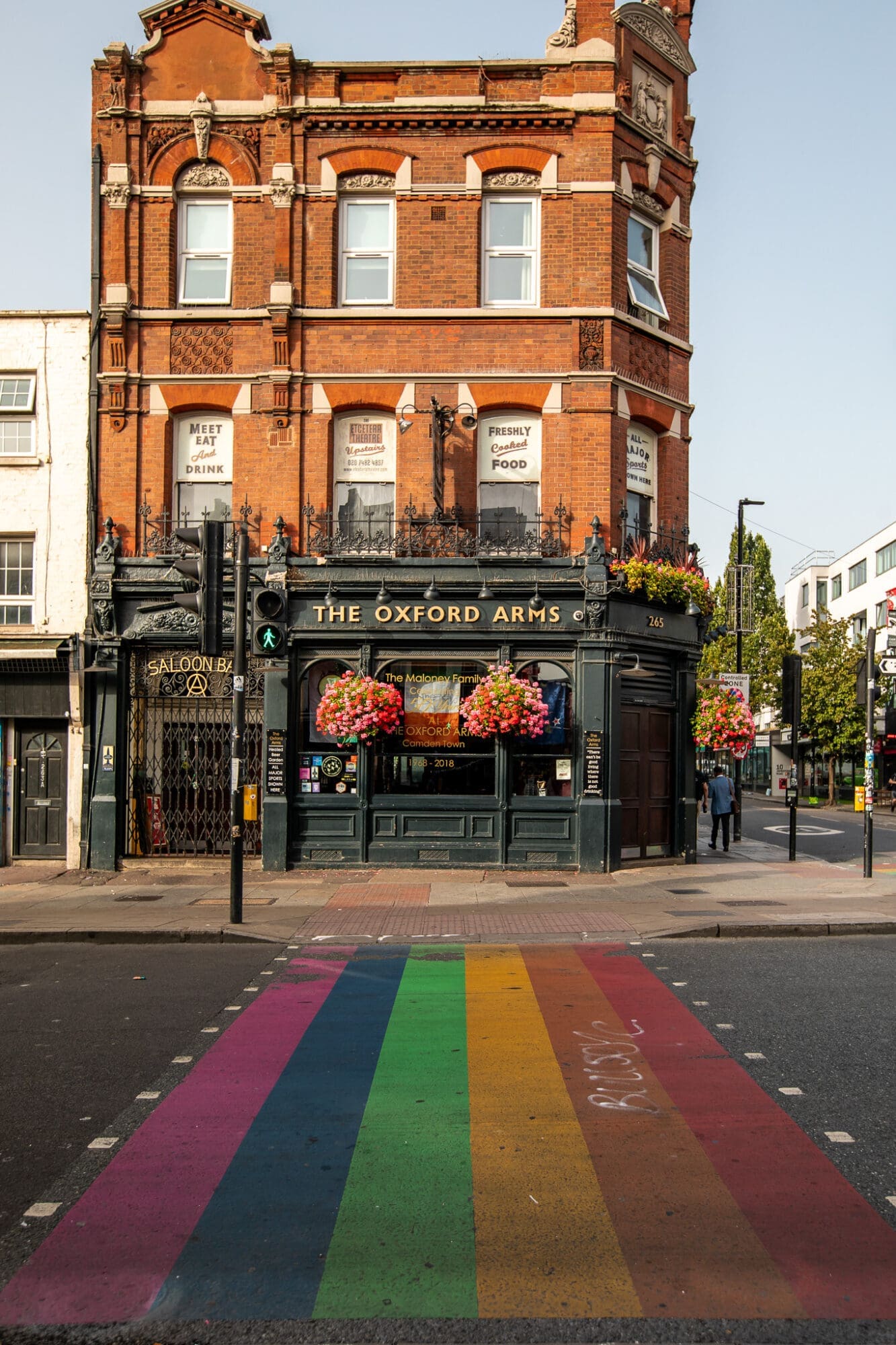 The Oxford Arms, a black pub with colourful hanging flower baskets, behind a rainbow painted "pride" zebra crossing