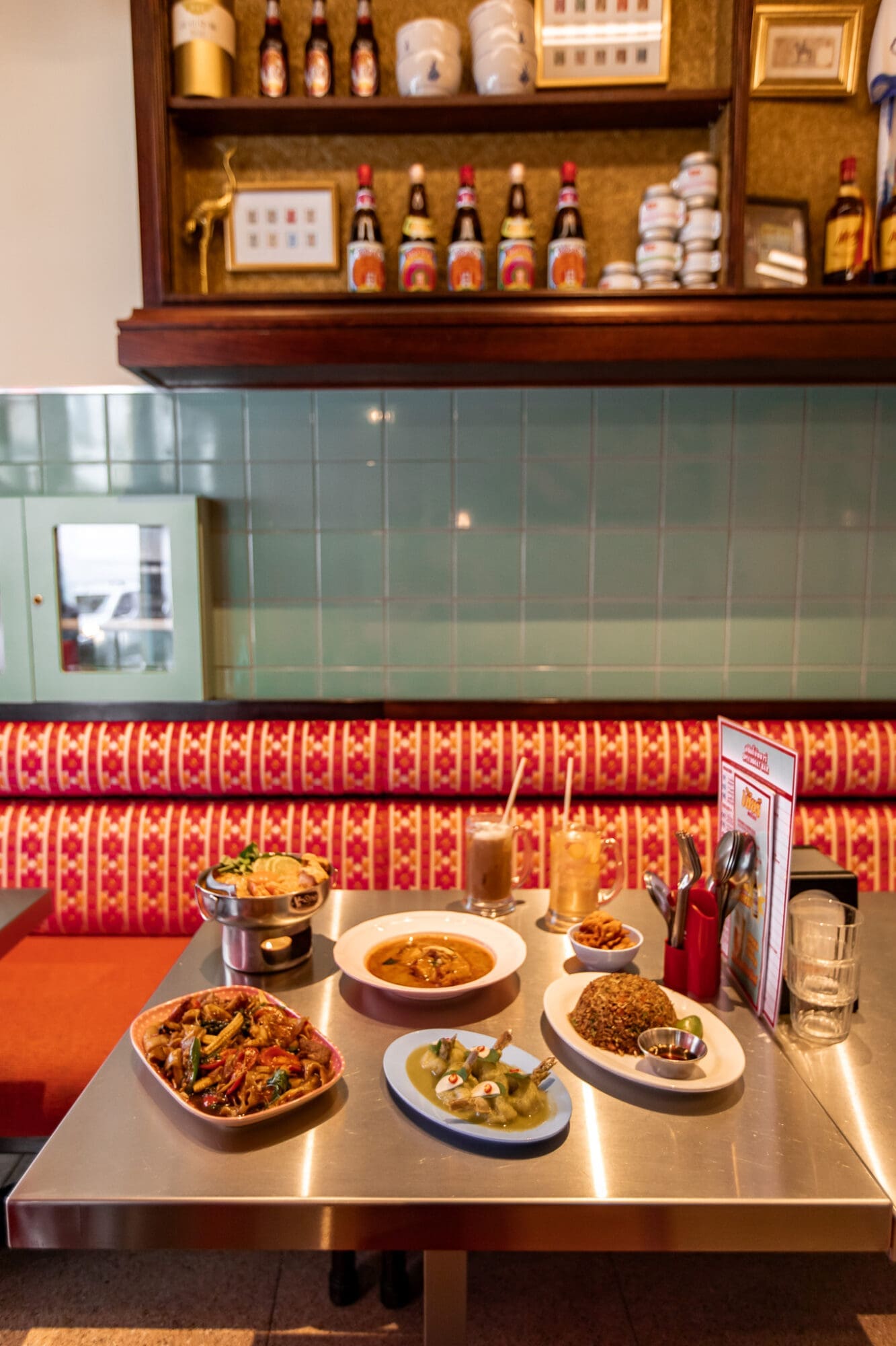A selection of dishes on a metal table against a red booth and green tiled wall at Speedboat Bar Thai Restaurant.