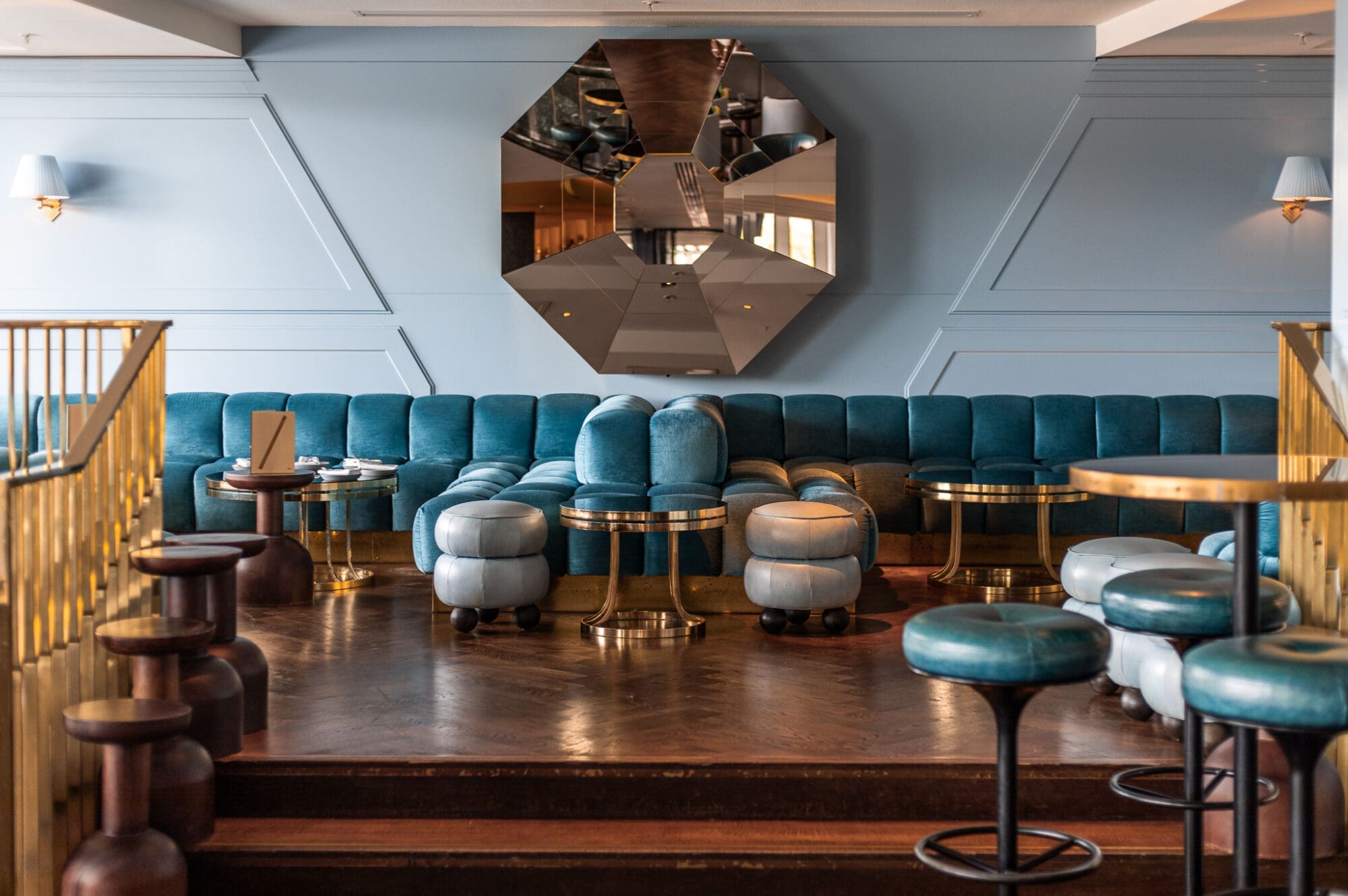 Plush blue sofa and chairs around tables against a blue wall and geometric mirror at Lyaness Cocktail Bar in London