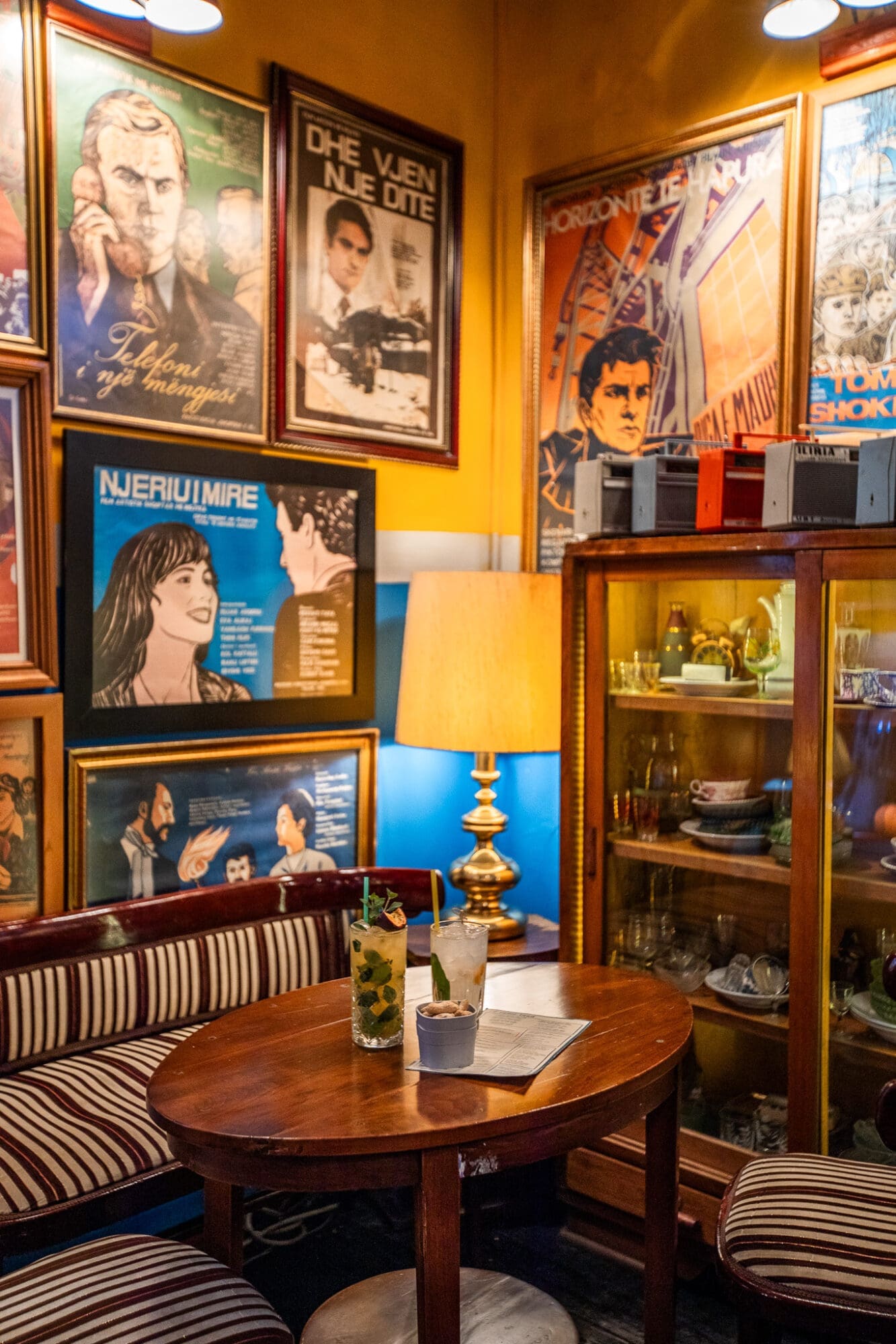 Two cocktails on a dark wood table surrounded by framed prints on the walls at a cocktail bar in Tirana, Albania