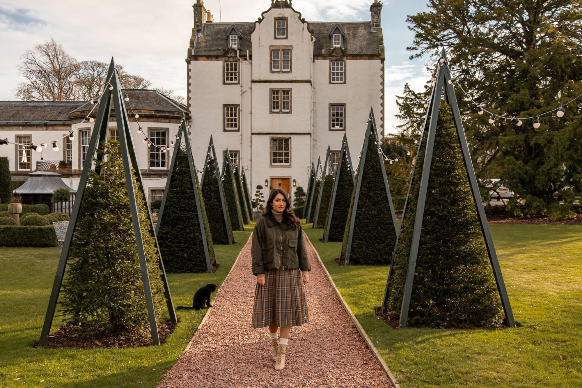 In the grounds of Prestonfield House Hotel in Edinburgh, Anoushka is wearing a checked skirt and walking on a path between triangular trees with the hotel in the background.