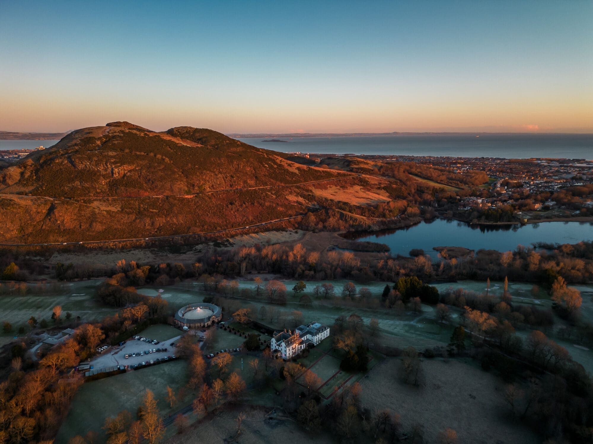 An aerial drone photograph taken over sunrise at Prestonfield House hotel with a view of Arthur's Seat and the sea in the background