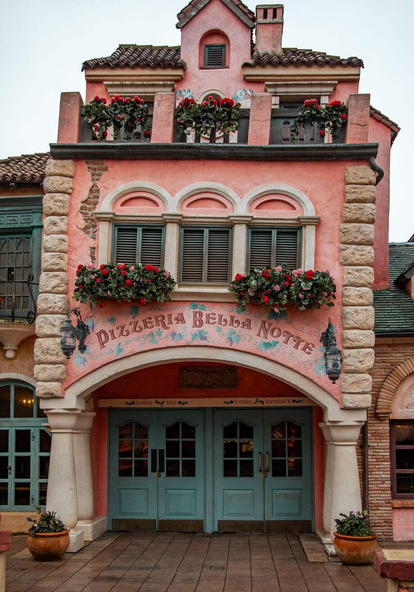 A pink, slightly off kilter restaurant facade with blue doors and flowers spilling out of the windows, of Pizzeria Bella Notte in Magic Kingdom Disneyland