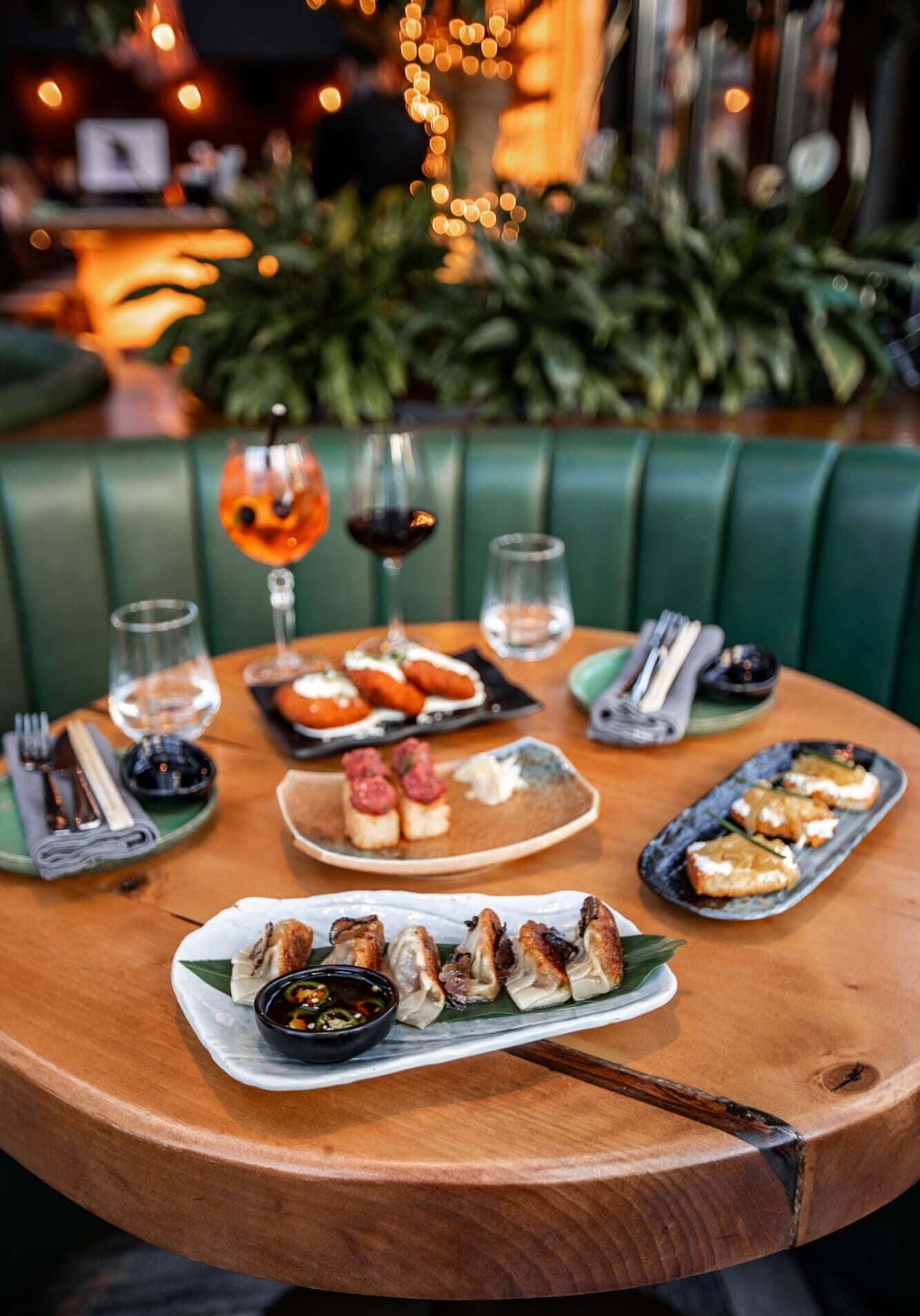 A selection of small plates including gyoza, crispy rice, and goats cheese crostini on a round wood table surrounded by a green leather booth and foliage.